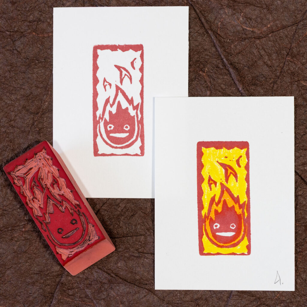 Calcifer (a stylized flame with two simple eyes and a mouth) printed in red on two pieces of rectangular white paper. The plain print is in the upper left. The print in the lower right has yellow and orange added with watercolor pencils. The eraser that made the prints is laying face-up at an angle in the lower left.