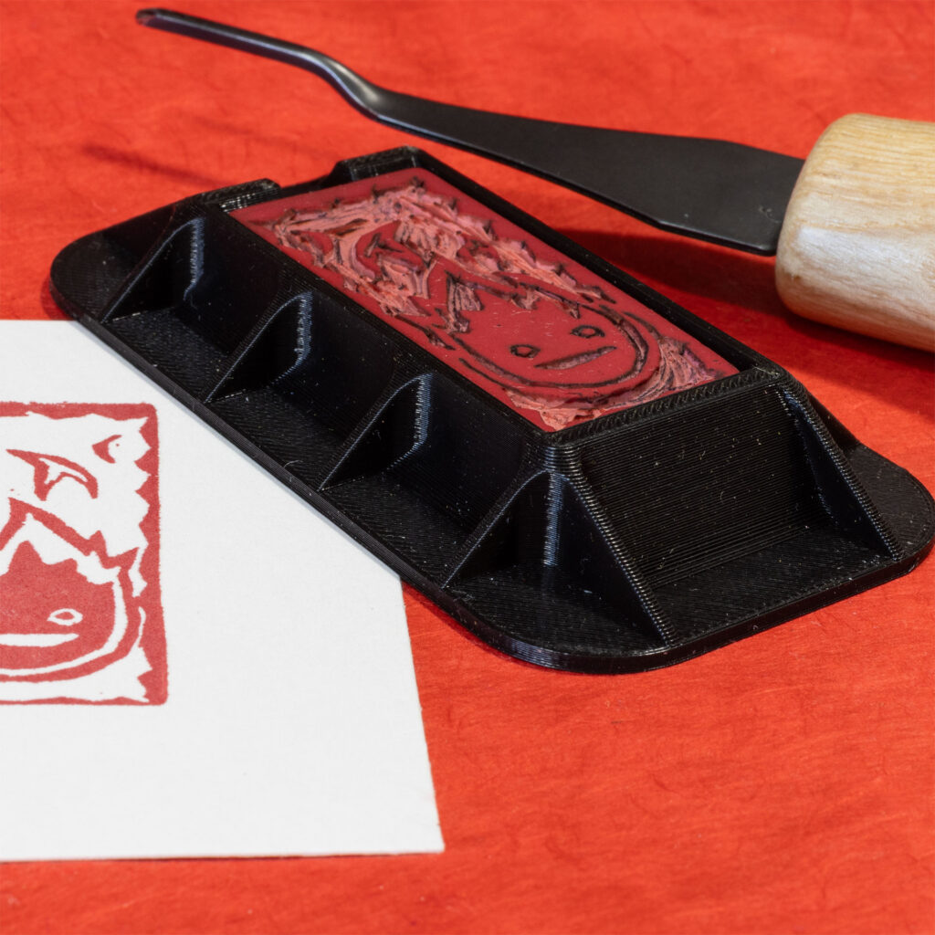 Angled view on a hand-carved Calcifer eraser (a stylized flame with two eyes and a mouth) in a black 3D-printed holder designed for carving erasers rests on a surface covered in red paper. A carving tool and part of a print made by the eraser are visible.