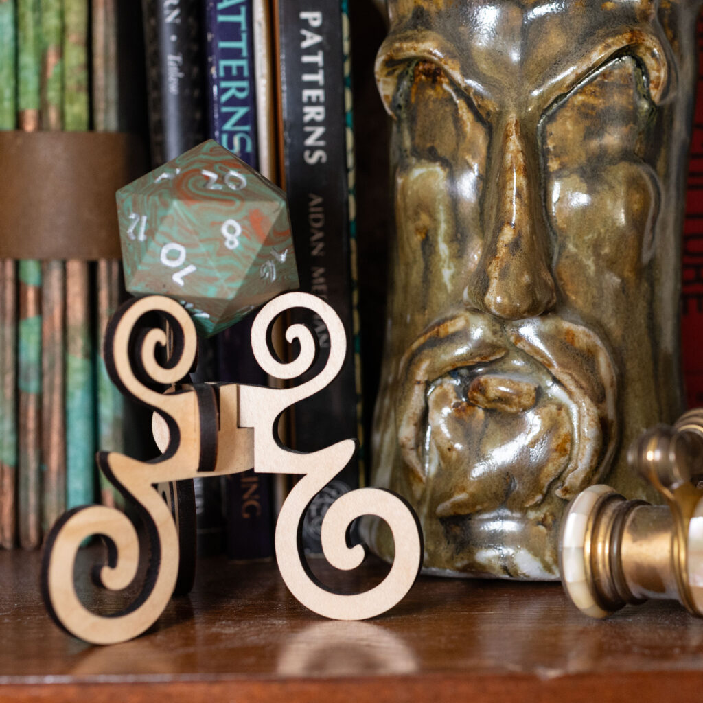 Side view of a large (42mm) 20-sided die (d20) swirled with teal an copper with the numbers painted in with metallic silver, resting on a three-legged stand with spiral elements made from maple. It is sitting on a bookshelf. Part of a ceramic container with a sculpted face and a pair of pearl-inlaid opera glasses are visible. Two of the books in the background have the word "Patterns" visible on the spine.