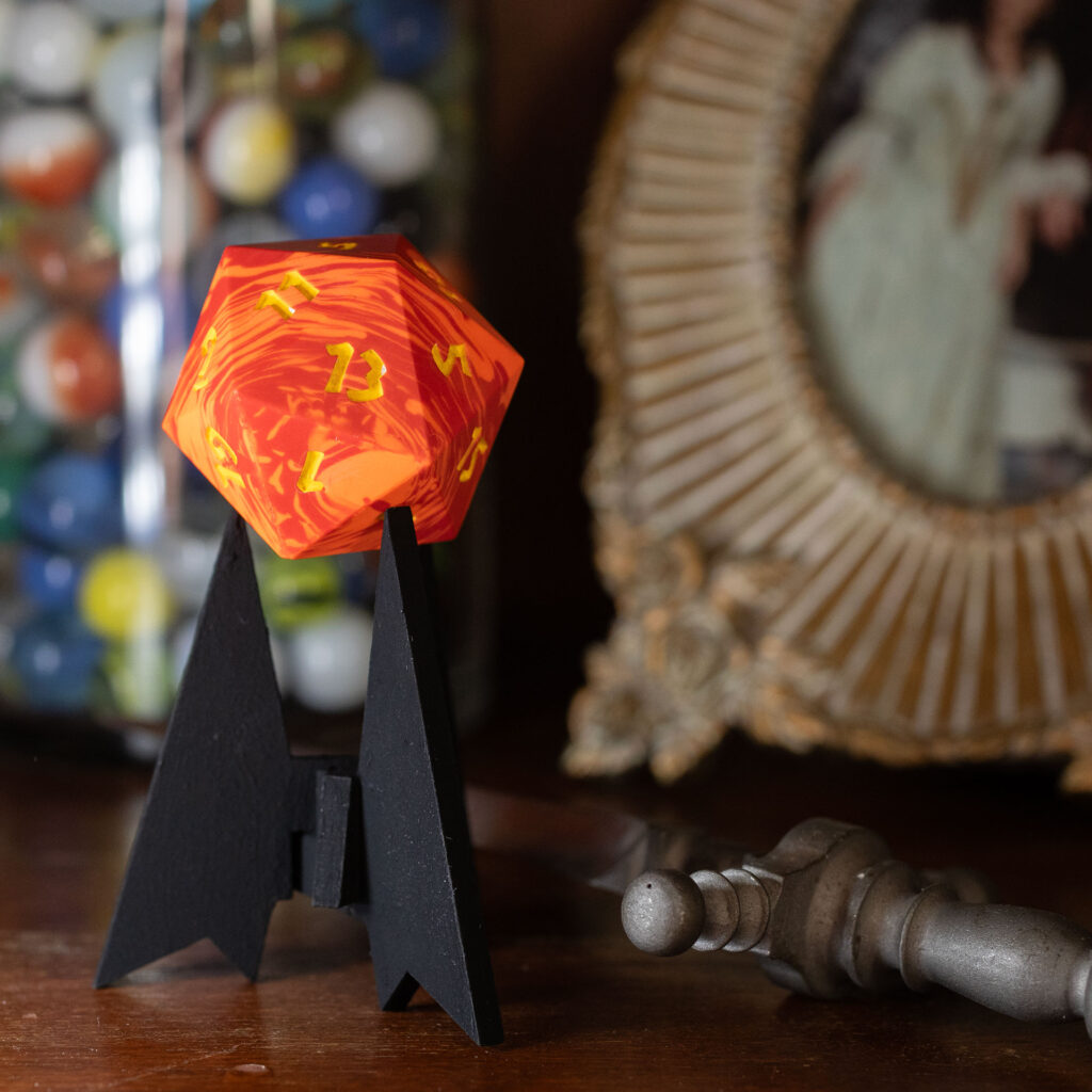 Side view of a large (42mm) 20-sided die (d20) swirled with two shades of yellow-orange with numbers painted in with bright yellow resting on a three-pointed stand in an unusually dark black. The 13 face is towards the view. It is sitting on a shelf. A jar of glass marbles, an ornately-framed picture, and stiletto are visible.