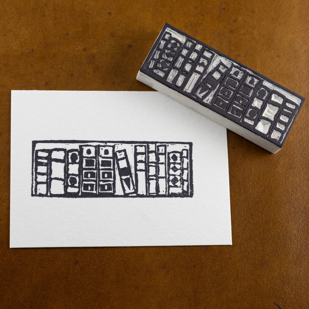 A white rectangle of paper imprinted with the image of a bookshelf with eleven books of different sizes. One book in the middle is tilted to the left within a larger gap. The hand-carved white blocky eraser that made the impression is laying face-up across the upper right corner. Both are on a brown surface.