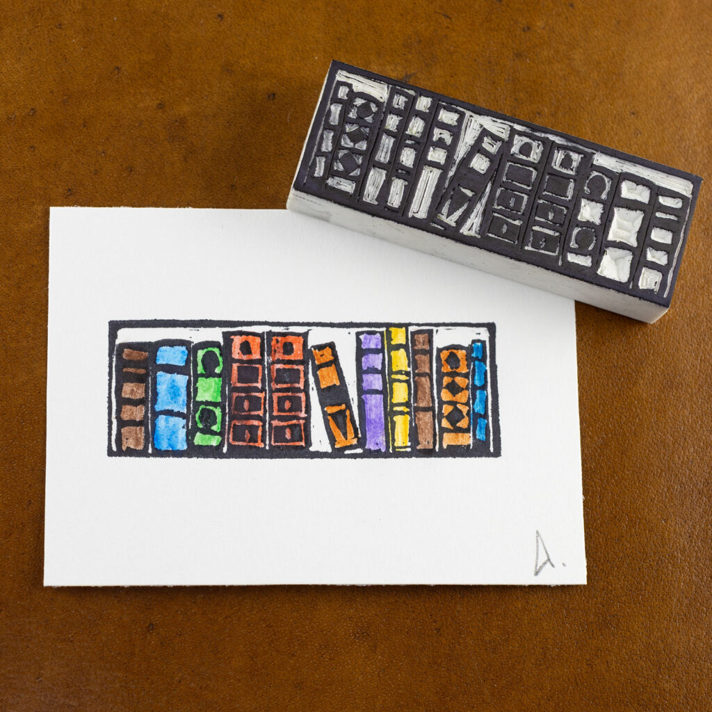 A white rectangle of paper imprinted with the image of a bookshelf with eleven books of different sizes. One book in the middle is tilted to the left within a larger gap. The books have been colored in with brown, blue, green, red, orange, purple, and yellow. The hand-carved white blocky eraser that made the impression is laying face-up across the upper right corner. Both are on a brown surface.