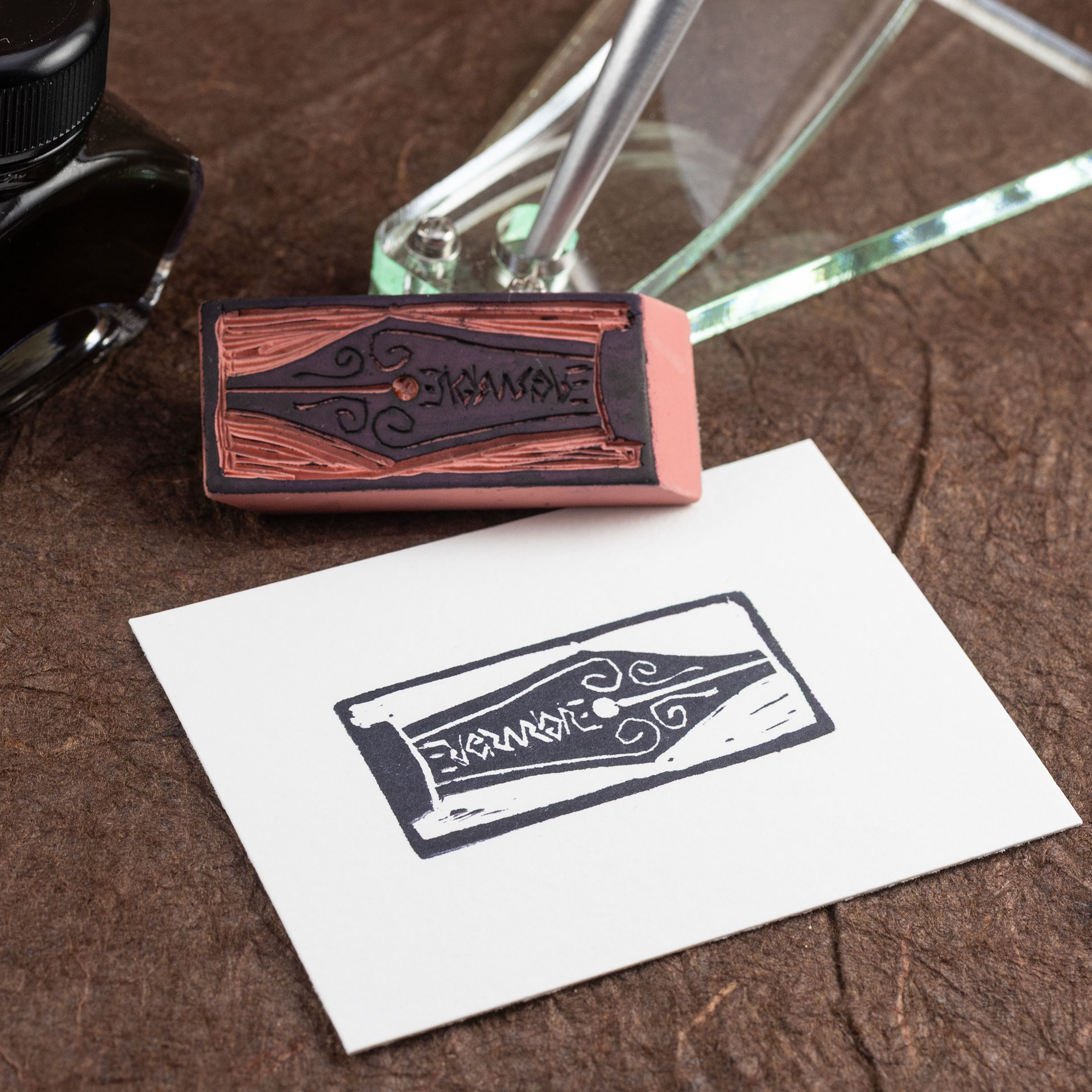 A white rectangle of paper imprinted with a stylized image of a fountain pen nib engraved with spirals and the Evermore ambigram sits on a surface covered with handmade dark brown paper. The hand-carved eraser that made the print is laying horizontally above the print, leaning against a translucent pen stand. Part of a bottle of writing ink is also visible in the background.