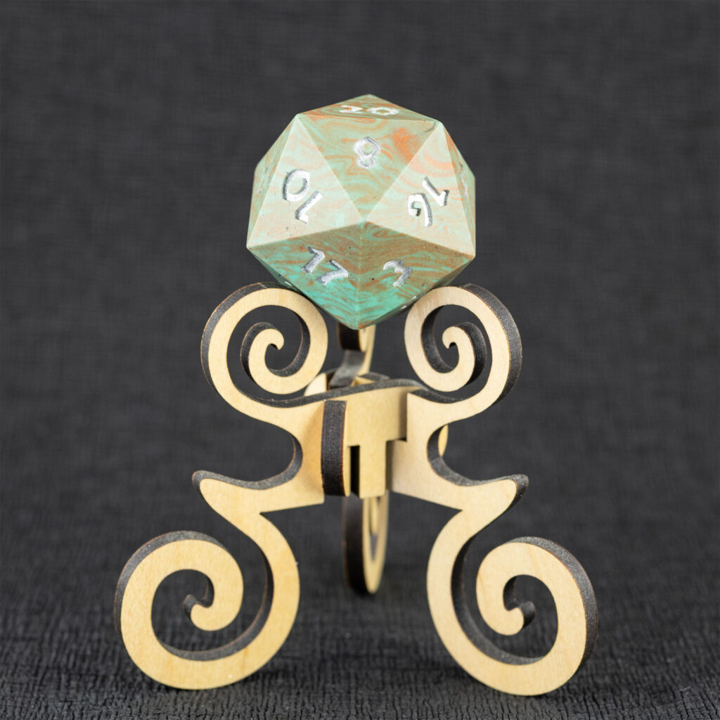 Three-quarters top-down view of a large (42mm) 20-sided die (d20) swirled with teal an copper with the numbers painted in with metallic silver, resting on a three-legged stand with spiral elements made from maple. The 20 side is facing up, with the 2, 8, 10, 12, 14, 16, and 17 faces visible. The background is a black creped paper (chirimen).