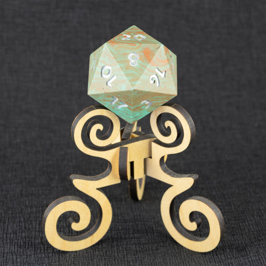 Three-quarters top-down view of a large (42mm) 20-sided die (d20) swirled with teal an copper with the numbers painted in with metallic silver, resting on a three-legged stand with spiral elements made from maple. The 20 side is facing up, with the 2, 8, 10, 12, 14, 16, and 17 faces visible. The background is a black creped paper (chirimen).