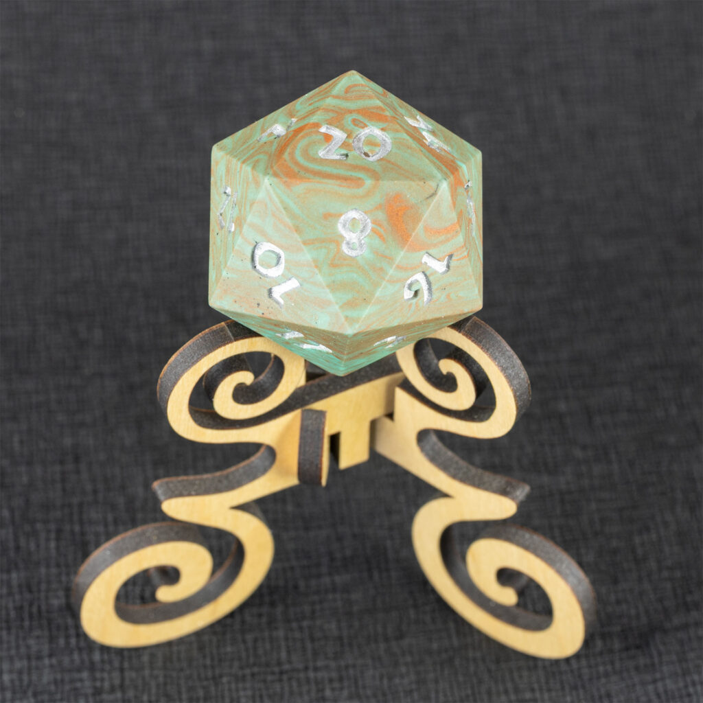 Three-quarters top-down view of a large (42mm) 20-sided die (d20) swirled with teal an copper with the numbers painted in with metallic silver, resting on a three-legged stand with spiral elements made from maple. The 20 side is facing up, with the 2, 8, 10, 12, 14, 16, and 17 faces visible. The background is a black creped paper (chirimen).