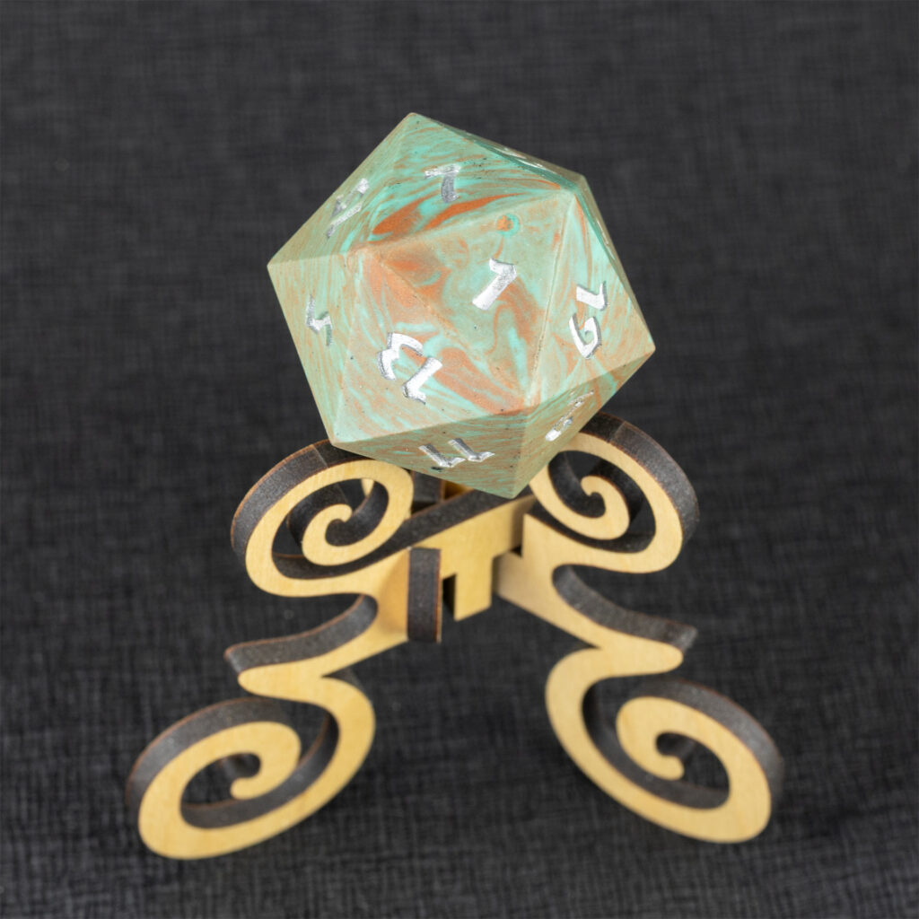 Three-quarters top-down view of a large (42mm) 20-sided die (d20) swirled with teal an copper with the numbers painted in with metallic silver, resting on a three-legged stand with spiral elements made from maple. The 7 side is facing up, with the 5, 9, 11, 13, 15, and 19 faces visible. The background is a black creped paper (chirimen).