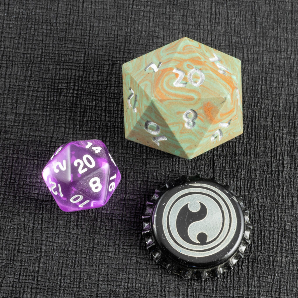 Top-down view of a large (42mm) 20-sided die (d20) swirled with teal an copper with the numbers painted in with metallic silver, a standard-sized translucent purple 20-sided die, and a bottle cap engraved with a Celtic spiral design resting on a sheet of black creped paper (chirimen).