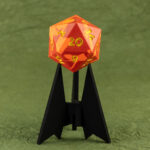 Side view of a large (42mm) 20-sided die (d20) swirled with two shades of yellow-orange with numbers painted in with bright yellow resting on a three-pointed stand in an unusually dark black. The 20 face is towards the viewer. The background is a mossy green crinkled paper (momigami).