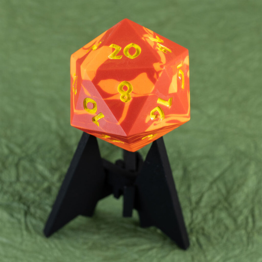 Three-quarters top-down view of a large (42mm) 20-sided die (d20) swirled with two shades of yellow-orange with numbers painted in with bright yellow resting on a three-pointed stand in an unusually dark black. The 20 face is pointing up and the 8 is facing the viewer. The background is a mossy green crinkled paper (momigami).
