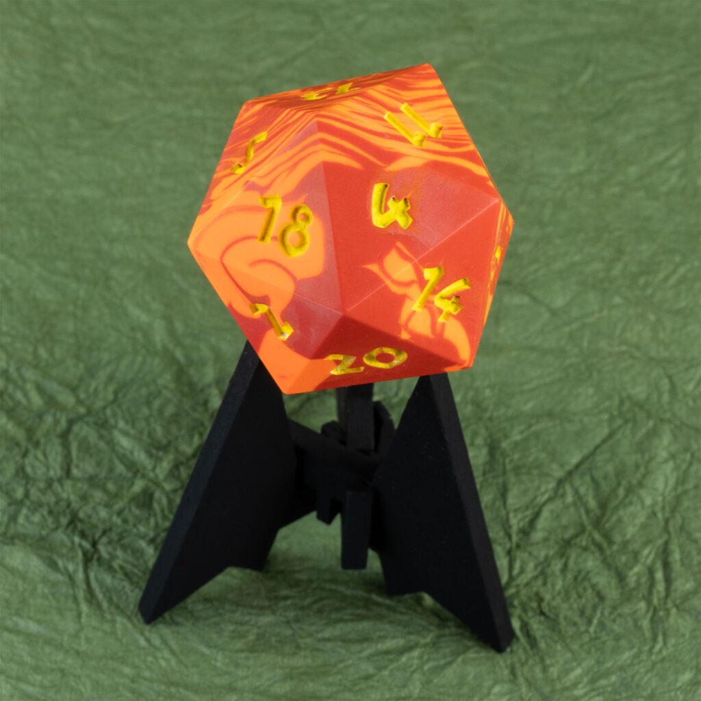Three-quarters top-down view of a large (42mm) 20-sided die (d20) swirled with two shades of yellow-orange with numbers painted in with bright yellow resting on a three-pointed stand in an unusually dark black. The 13 face is pointing up, with the 2, 4, 5, 11, 14,, and 20 faces are visible. The background is a mossy green crinkled paper (momigami).