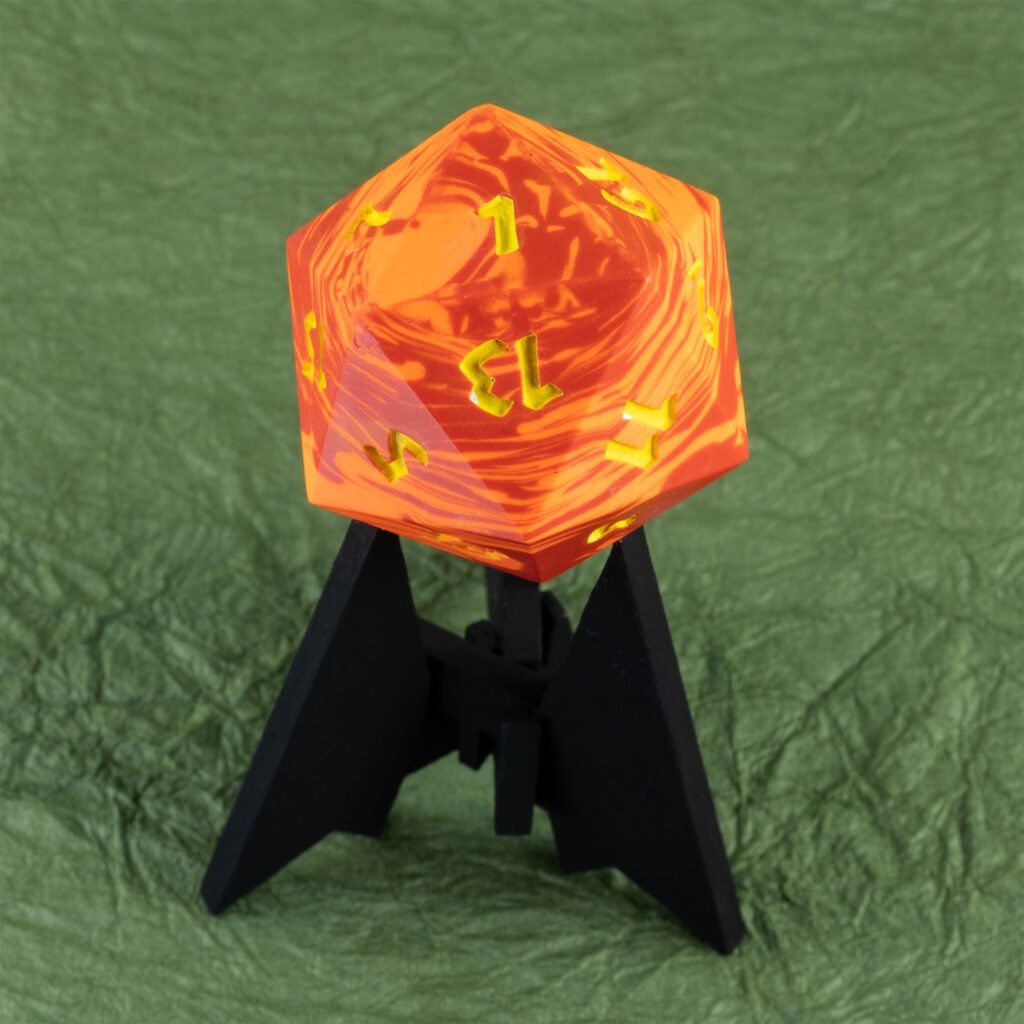 Three-quarters top-down view of a large (42mm) 20-sided die (d20) swirled with two shades of yellow-orange with numbers painted in with bright yellow resting on a three-pointed stand in an unusually dark black. The 1 face is pointing up and toward the viewer, the 13 face is upside down facing the viewer, and the 3, 5, 7, 9, 15, and 19 faces are visible. The background is a mossy green crinkled paper (momigami).
