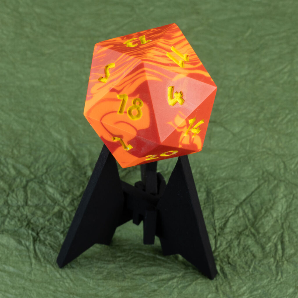 Three-quarters top-down view of a large (42mm) 20-sided die (d20) swirled with two shades of yellow-orange with numbers painted in with bright yellow resting on a three-pointed stand in an unusually dark black. The 13 face is pointing up, with the 2, 4, 5, 11, 14,, and 20 faces are visible. The background is a mossy green crinkled paper (momigami).