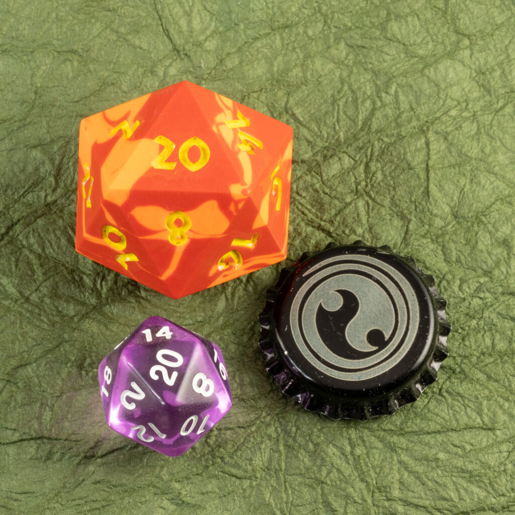 Top-down view of a large (42mm) 20-sided die (d20) swirled with two shades of yellow-orange with numbers painted in with bright yellow, a standard-sized translucent purple 20-sided die, and a bottle cap engraved with a Celtic spiral design resting on a sheet of crinkled moss-green paper (momigami).