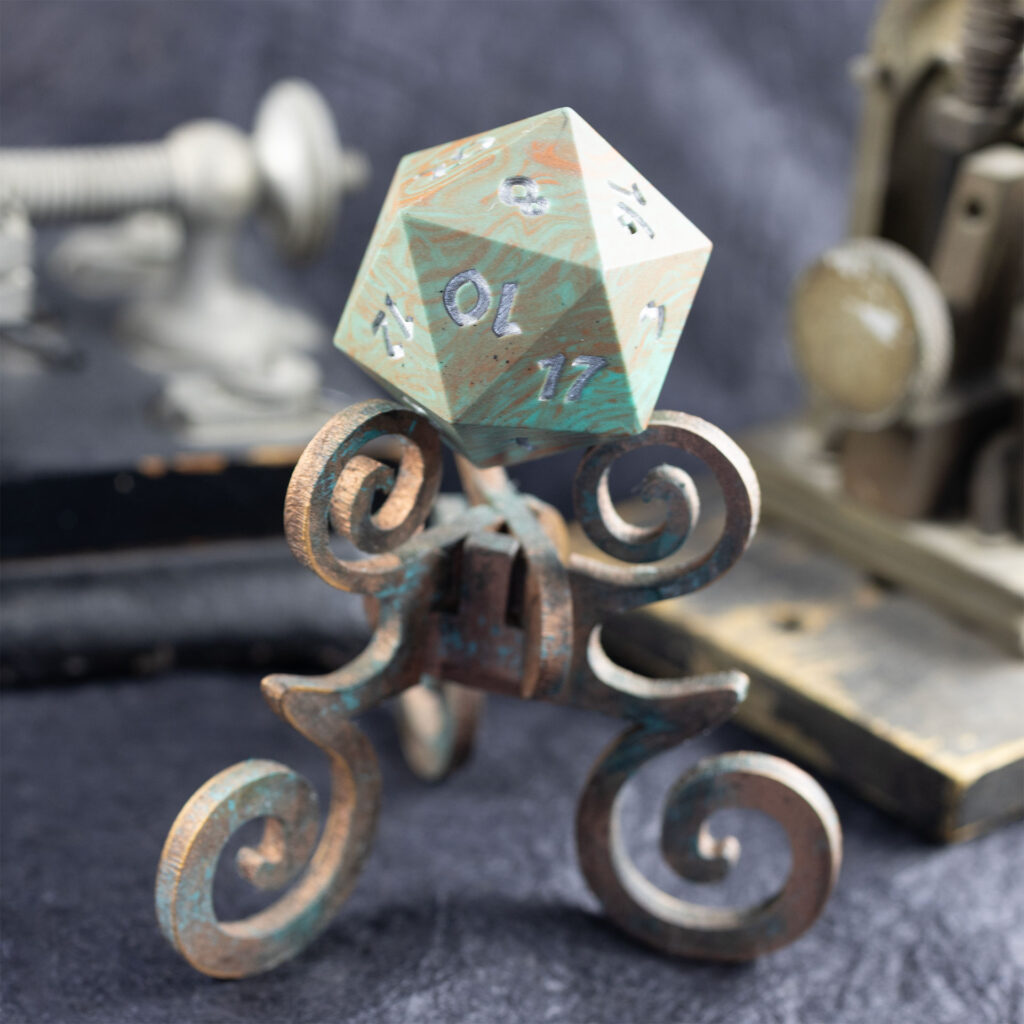 A large (42mm) 20-sided die (d20) swirled with teal an copper with the numbers painted in with metallic silver rests on a three-legged stand with spiral elements and finished with a faux copper patina. It sits on a textured black surface with some very old-looking electrical equipment out-of-focus in the background (parts of an old telegraph system).
